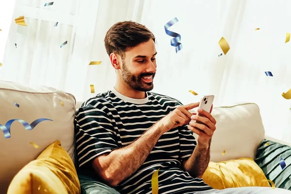 Young man sitting on a sofa surrounded by confetti while looking at a phone.